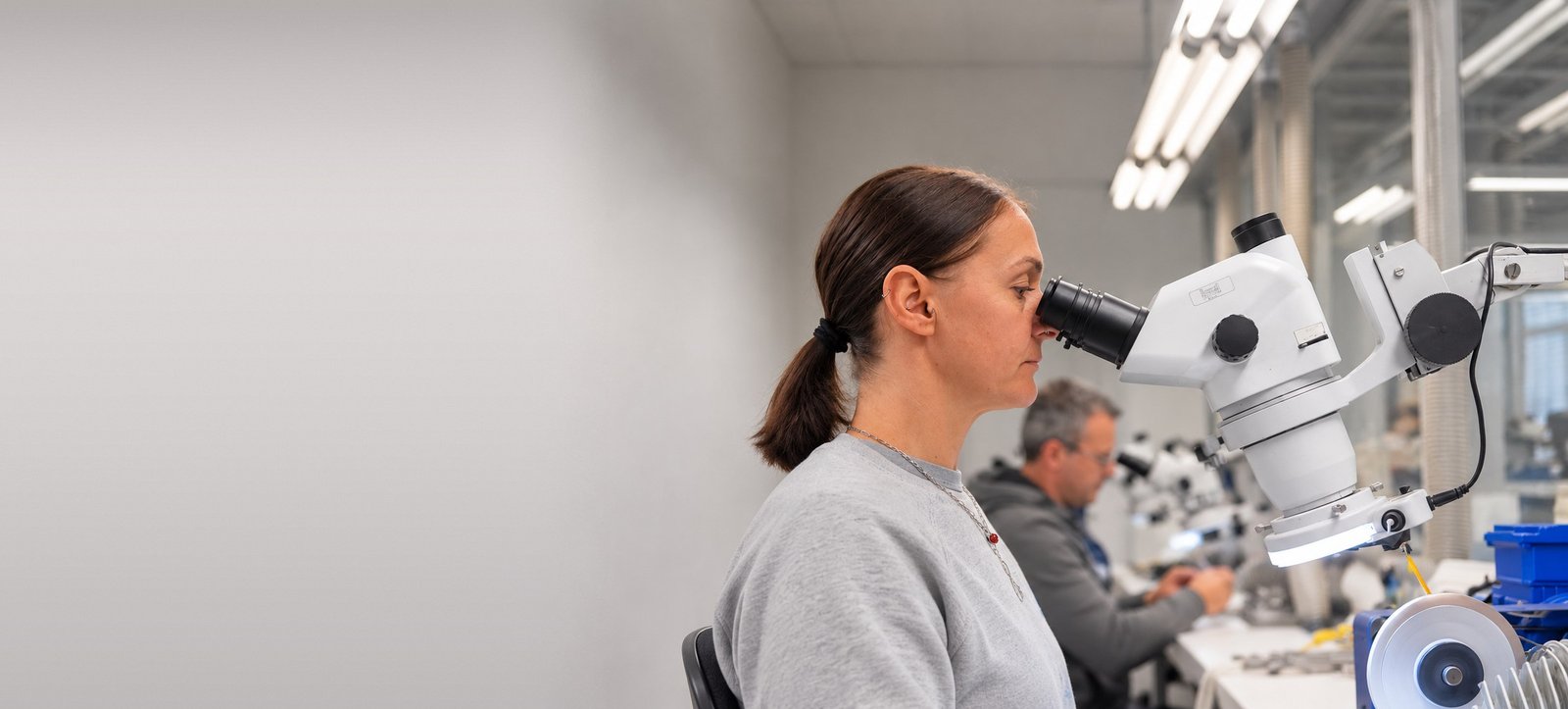 Mitarbeiterin prüft ein Instrument am Stereomikroskop, im Hintergrund ein Kollege am Arbeitsplatz
